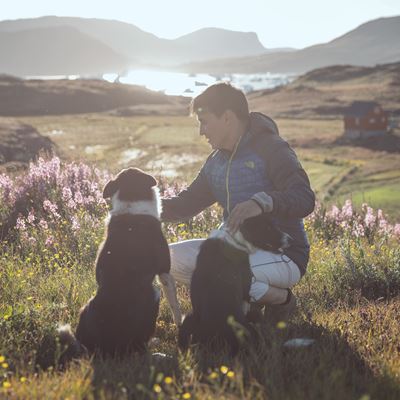 En mand sidder med sin hund udenfor i solskinsvejr med en flot udsigt til grønne bjerge.