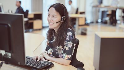 Mørkhåret kvinde sidder ved sit kontor foran sin computer og snakker tlf. med headset. 
