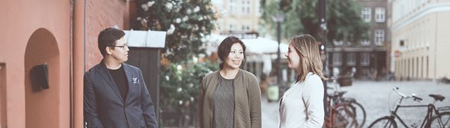 Tre unge mennesker står på gaden ved siden af en rød bygning. 