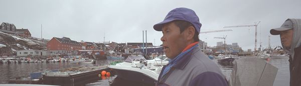 En mand med en kasket står udenfor i havnen med en båd baggrunden. 