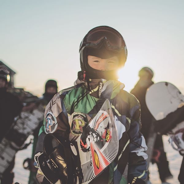 Barn iklædt snooboardtøj sammen med andre mennesker og en flot pist baggrund.
